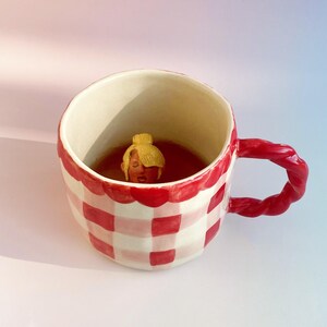 Può includere: Una tazza di gingham rossa e bianca con un manico a corda rosso. Una piccola figurina di ceramica di una donna con i capelli biondi è all'interno della tazza.