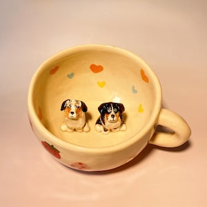 May include: A white ceramic mug with a handle, decorated with pink, blue and yellow hearts. Two small ceramic dog figurines are inside the mug, one with brown, black and white fur and the other with black and white fur.