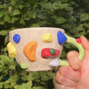 Pode incluir: Uma caneca de cerâmica bege com superfície texturizada, decorada com várias formas coloridas e elevadas de agarras de escalada em azul, amarelo, laranja, vermelho e branco. A caneca possui uma alça verde-clara com um pequeno ponto vermelho.