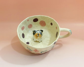 May include: A white ceramic mug with a handle, decorated with pink and purple polka dots and floral designs. A small ceramic dog figurine is sitting inside the mug.
