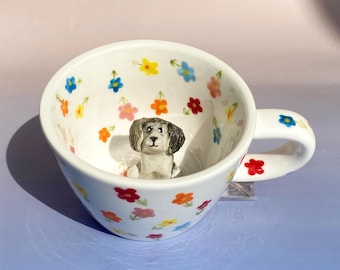 Puede incluir: Una taza de té de cerámica blanca con una pequeña figura de perro en su interior. La taza está decorada con un patrón floral colorido.