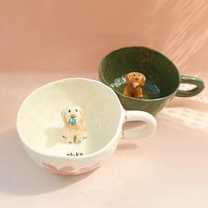 Może przedstawiać: Dwa ceramiczne kubki z figurkami psów w środku. Jeden kubek jest kremowy z różowymi kwiatowymi akcentami i psem trzymającym niebieską piłkę. Drugi kubek jest zielony z brązową figurką psa. Oba kubki mają uchwyty.