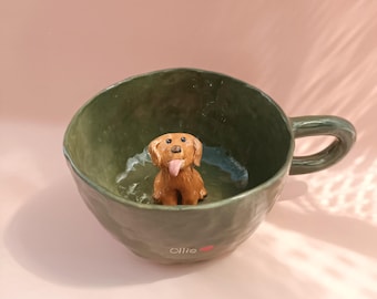 Puede incluir: Una taza de cerámica verde con una figurita de perro marrón en su interior. La taza tiene la palabra "ollie" escrita en la parte inferior.