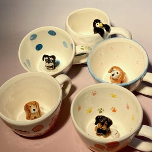 May include: Five ceramic mugs with different animal figurines inside. The mugs have various patterns, including blue polka dots, paw prints, and a light blue background with pink dots. The figurines include a penguin, a dachshund, a beagle, a schnauzer, and a golden retriever.