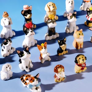 以下が含まれることがあります： さまざまな犬種とポーズのセラミック犬の置物のコレクション。犬は白、黒、茶色、オレンジなど、さまざまな色で塗装されています。帽子、リボン、スカーフを身に着けているものもあります。背景は単色の水色です。