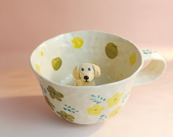 Puede incluir: Una taza de cerámica blanca con un patrón floral y lunares amarillos y verdes. Una pequeña figurita de perro de cerámica está dentro de la taza.