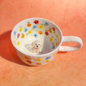 May include: White ceramic mug with a small white dog figurine inside. The mug is decorated with a floral pattern of red, yellow, blue, and orange flowers.