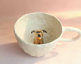 May include: A white ceramic teacup with a textured exterior and a brown and black dog figurine inside. The dog is holding a small orange ball in its mouth.