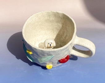 May include: White ceramic mug with a small white dog figurine inside. The mug has a handle and is decorated with colorful, abstract shapes.
