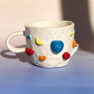 Può includere: Tazza in ceramica bianca con superficie testurizzata e forme 3D colorate, tra cui rosso, giallo, blu, arancione e verde.