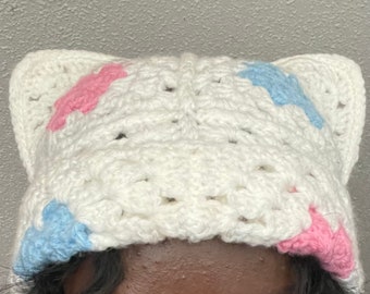 May include: A handmade white crochet hat with cat ears. The hat features pink and blue accents on the ears. The hat is designed with a textured stitch pattern and is suitable for all genders.