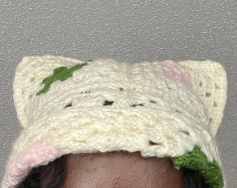 May include: Cream-colored crochet hat with cat ear design. The hat features green and pink floral accents. The hat is handmade and has a textured appearance. The hat is worn by a person with dark hair.