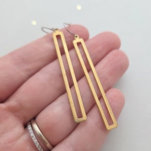 May include: A pair of long, rectangular gold-toned earrings featuring an open-frame design. Each earring consists of a thin, vertical metal bar with a hollow center, suspended from a simple silver-toned hook wire for attachment to the ear.