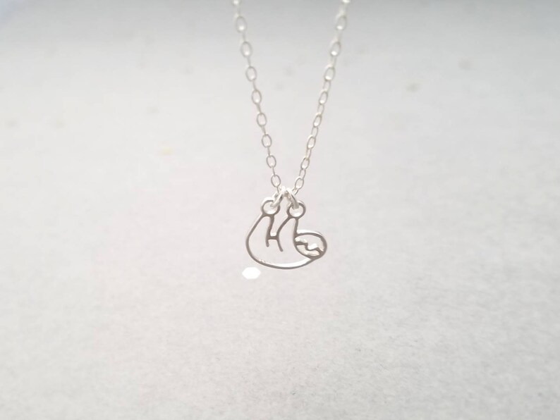 May include: A silver necklace with a delicate, hanging sloth charm. The sloth is depicted in a simple, minimalist style.