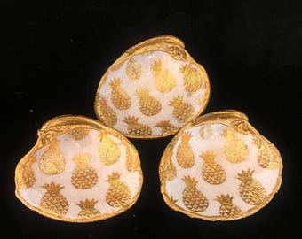 May include: Three decorative oyster shells with a white interior and gold pineapple pattern. The shells have a gold trim and are arranged on a black surface. The shells are likely used for serving or display.