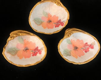 May include: Three decorative oyster shells with a gold rim and painted floral designs. Each shell features a central orange flower with pink and red accents on a white background. The shells are arranged on a black surface.