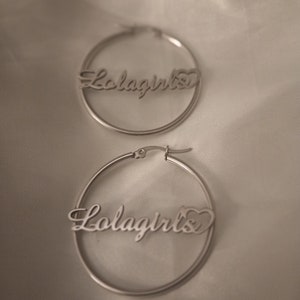 Puede incluir: Un par de pendientes de aro de plata con la palabra "Lolagirls" y un s&iacute;mbolo de coraz&oacute;n.