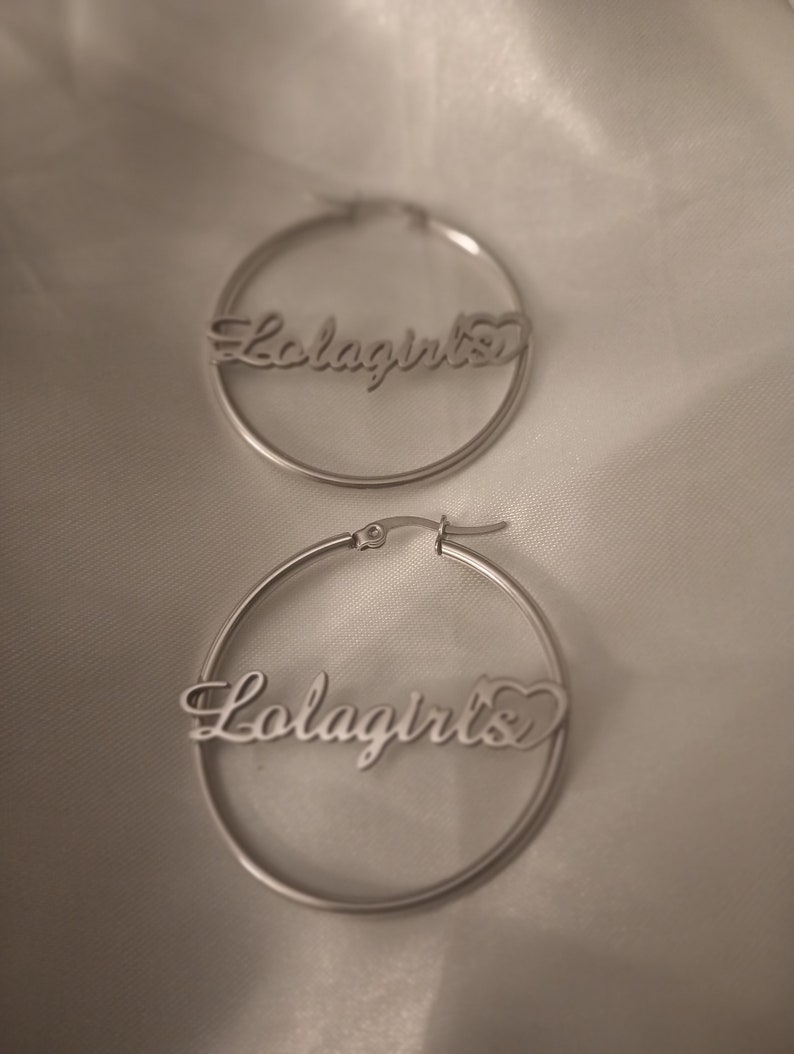 Puede incluir: Un par de pendientes de aro de plata con la palabra "Lolagirls" y un s&iacute;mbolo de coraz&oacute;n.