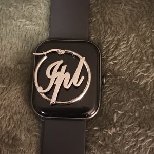 Puede incluir: Un pendiente de aro plateado con el nombre "Jhl" inscrito en letra cursiva. El pendiente se muestra en un reloj inteligente negro.