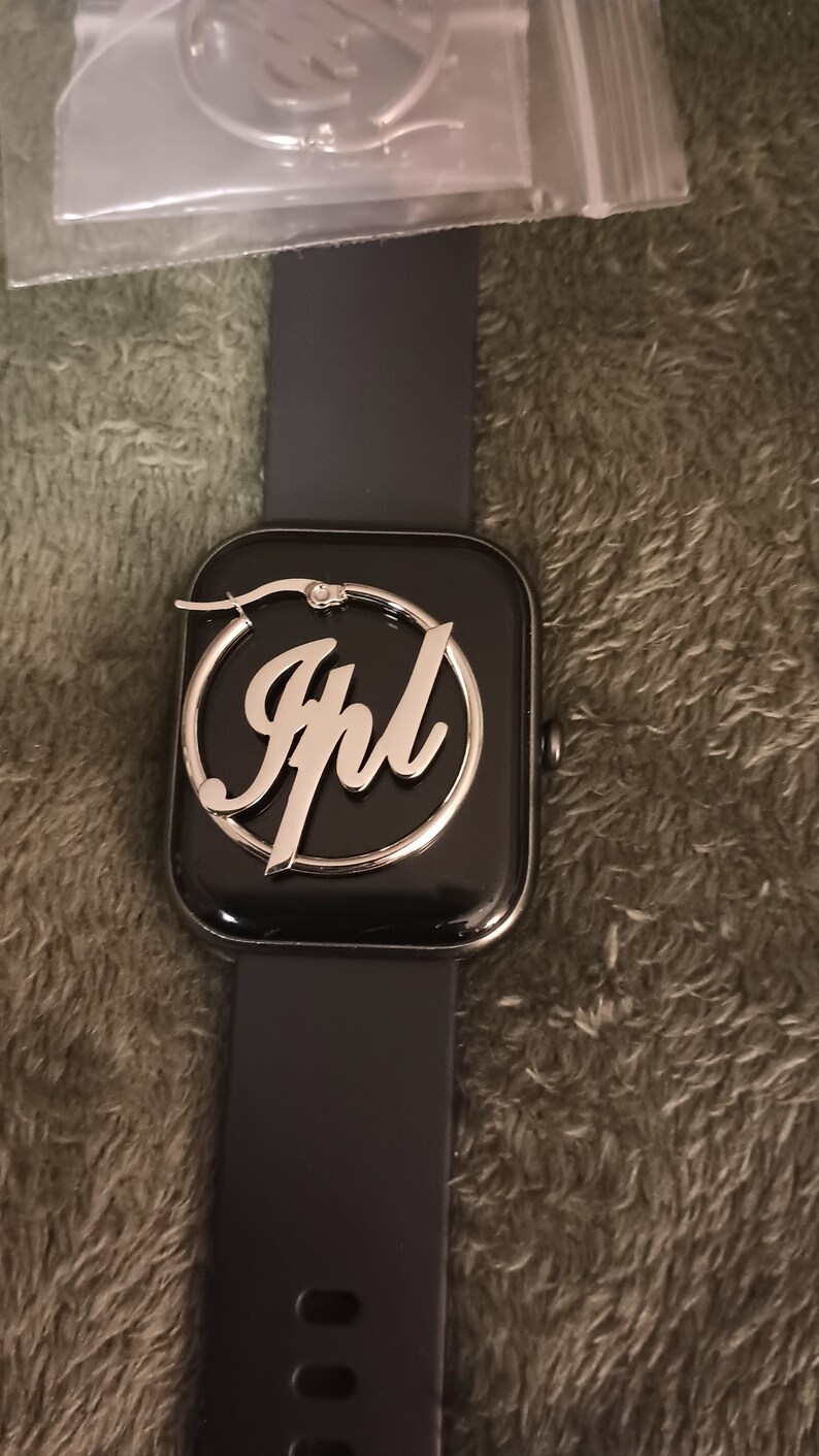 Puede incluir: Un pendiente de aro plateado con el nombre "Jhl" inscrito en letra cursiva. El pendiente se muestra en un reloj inteligente negro.