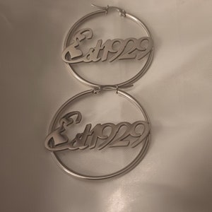 Puede incluir: Un par de pendientes de aro de plata con el texto "Est 1929" inscrito en cada aro.