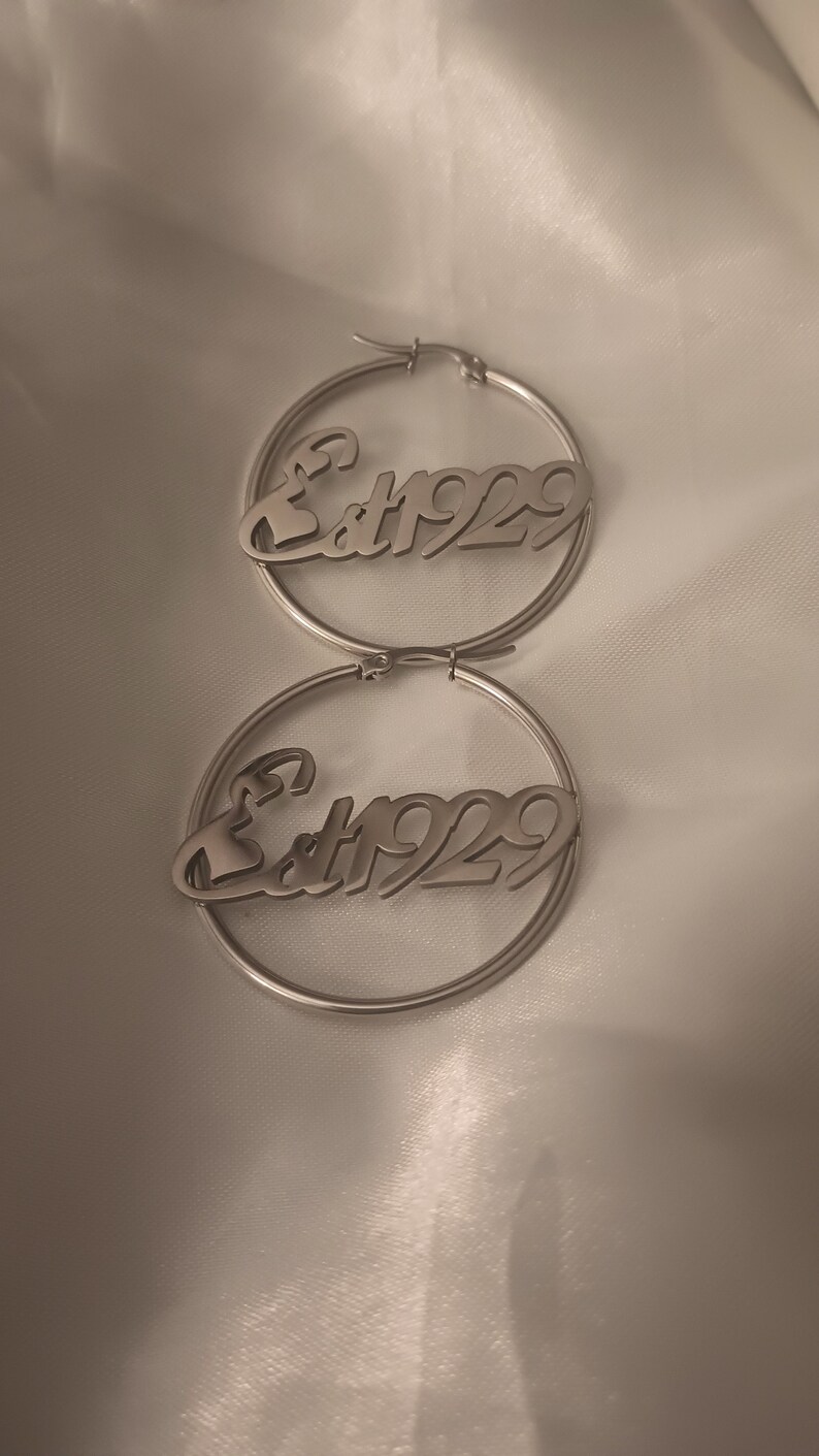 Puede incluir: Un par de pendientes de aro de plata con el texto "Est 1929" inscrito en cada aro.