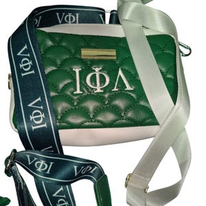 Op de afbeelding: Een groene en witte quilted crossbody tas met een witte band en een groene band met de letters "VΦI" herhaald. De tas heeft een gouden rits en de letters "IΦΛ" geborduurd op de voorkant.