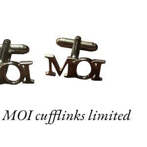 Op de afbeelding: Een paar zilveren manchetknopen met het woord "MOI" op elk. De manchetknopen zijn gemaakt door "MOI cufflinks limited".