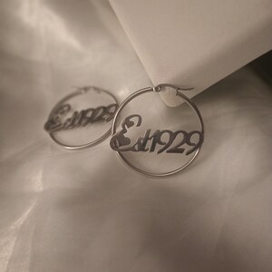 Puede incluir: Un par de pendientes de aro de plata con el texto "Est. 1929" inscrito en ellos.
