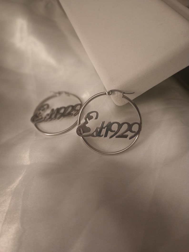 Puede incluir: Un par de pendientes de aro de plata con el texto "Est. 1929" inscrito en ellos.