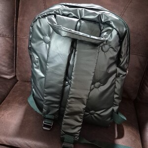 Peut inclure: Un sac &agrave; dos matelass&eacute; vert fonc&eacute; avec des bretelles r&eacute;glables. Le sac &agrave; dos a une finition brillante, presque m&eacute;tallique.
