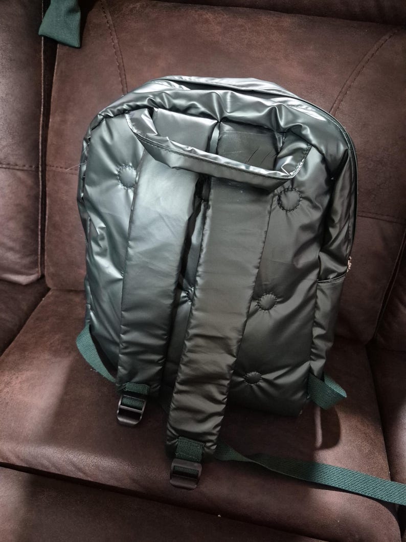Peut inclure: Un sac &agrave; dos matelass&eacute; vert fonc&eacute; avec des bretelles r&eacute;glables. Le sac &agrave; dos a une finition brillante, presque m&eacute;tallique.