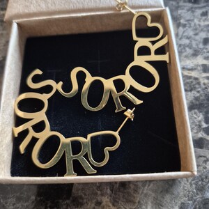 Puede incluir: Pendientes de aro de oro con la palabra "SOROR" escrita en un dise&ntilde;o curvo. Los pendientes est&aacute;n en una peque&ntilde;a caja.