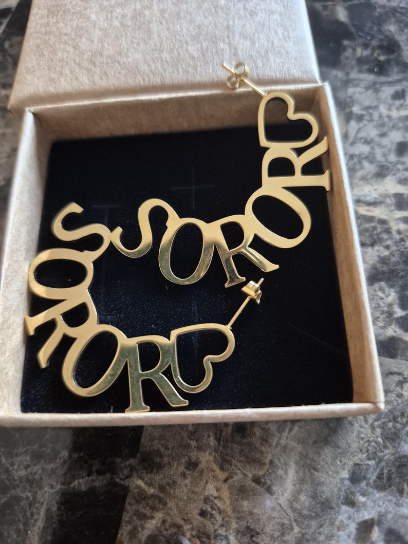 Puede incluir: Pendientes de aro de oro con la palabra "SOROR" escrita en un dise&ntilde;o curvo. Los pendientes est&aacute;n en una peque&ntilde;a caja.