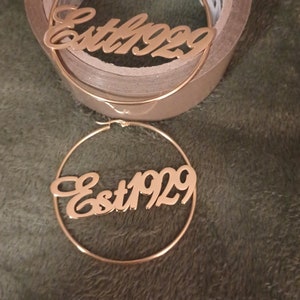 Puede incluir: Un par de pendientes de aro de oro con el texto "Est 1929" inscrito en la parte delantera.