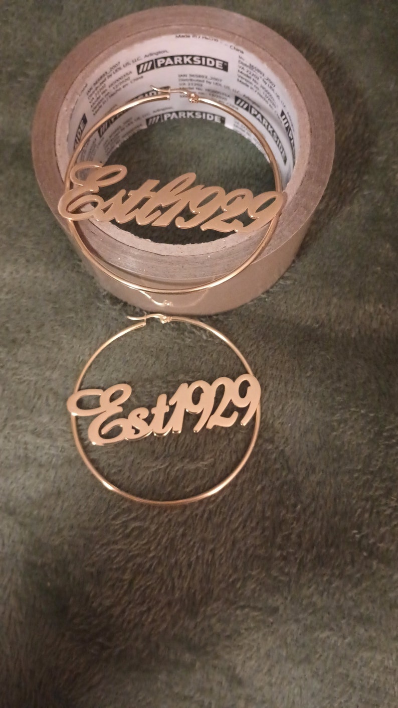 Puede incluir: Un par de pendientes de aro de oro con el texto "Est 1929" inscrito en la parte delantera.