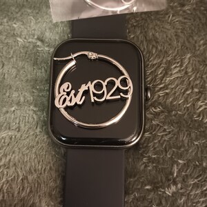 Puede incluir: Un pendiente de aro plateado con el texto "Est 1929" en un reloj inteligente negro.