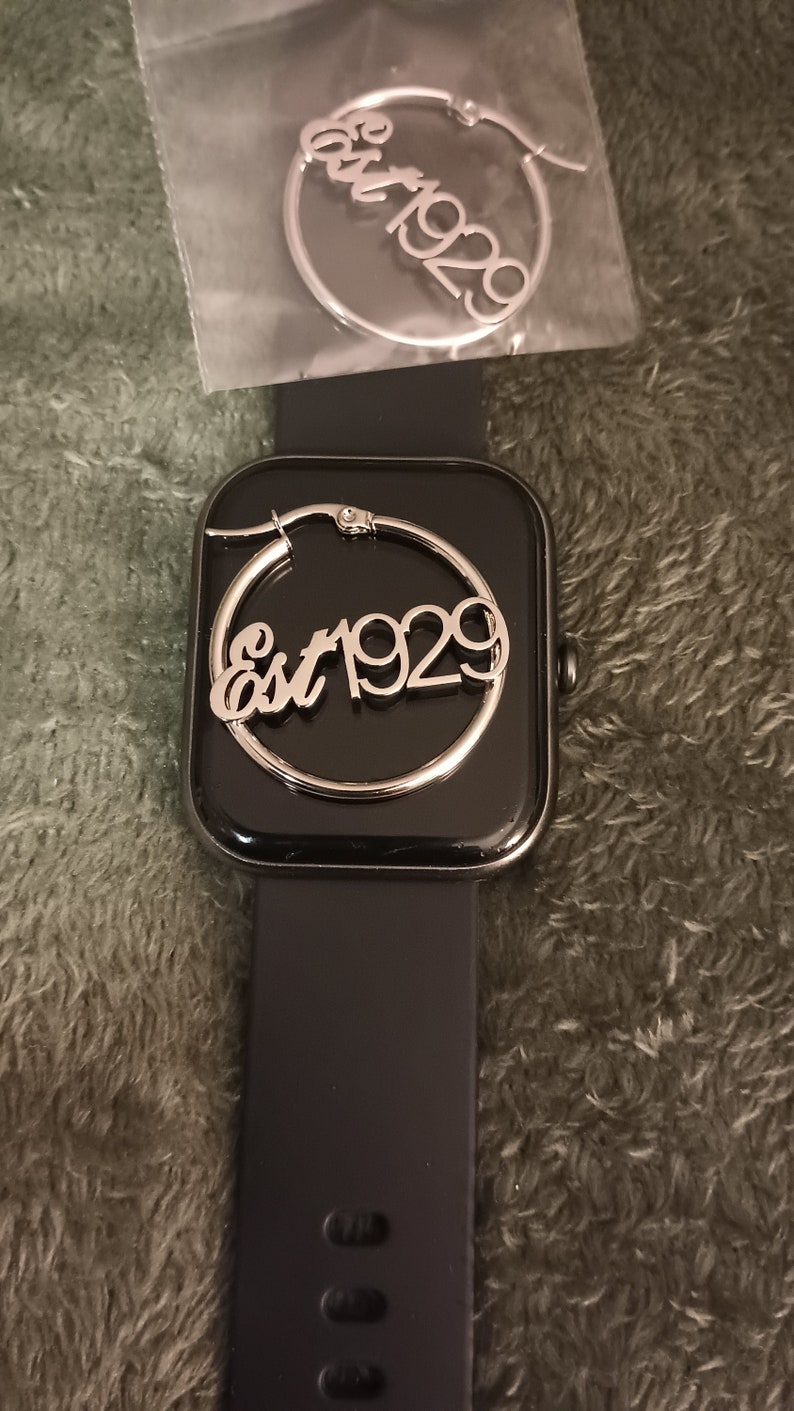 Puede incluir: Un pendiente de aro plateado con el texto "Est 1929" en un reloj inteligente negro.