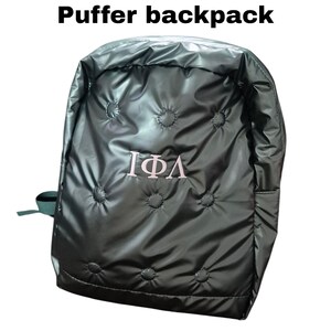 Peut inclure: Un sac &agrave; dos matelass&eacute; vert fonc&eacute; avec un logo brod&eacute; en blanc repr&eacute;sentant les lettres grecques "I&Phi;A".