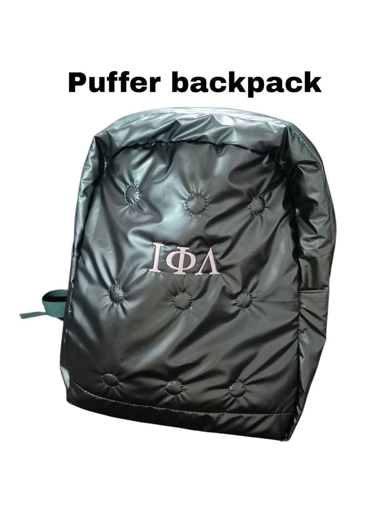 Peut inclure: Un sac &agrave; dos matelass&eacute; vert fonc&eacute; avec un logo brod&eacute; en blanc repr&eacute;sentant les lettres grecques "I&Phi;A".