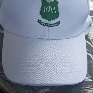 Puede incluir: Gorra de béisbol blanca con visera curva. El emblema verde y blanco muestra las letras griegas IΦΛ y el texto "IOTA PHI LAMBDA 29". Una pequeña flor blanca está adherida al emblema.