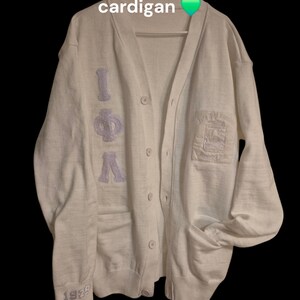 May include: White cardigan with a purple embroidered "IPL" on the left side and a small embroidered patch on the right side. The cardigan has a button closure and two pockets. The text "1929" is embroidered on the left sleeve.