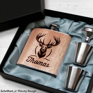Könnte beinhalten: Eine Holzflasche mit einem Hirsch-Design und dem eingravierten Namen "Thomas". Die Flasche befindet sich in einer schwarzen Box mit blauer Satin-Auskleidung. Die Box enthält außerdem zwei Schnapsgläser und einen Trichter.