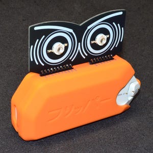 Puede incluir: Un dispositivo electr&oacute;nico naranja con un componente en blanco y negro. El componente negro tiene un dise&ntilde;o que se asemeja a ojos. El dispositivo tiene la palabra japonesa "Flipper" impresa.