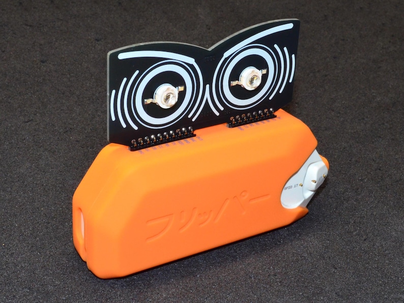 Puede incluir: Un dispositivo electr&oacute;nico naranja con un componente en blanco y negro. El componente negro tiene un dise&ntilde;o que se asemeja a ojos. El dispositivo tiene la palabra japonesa "Flipper" impresa.