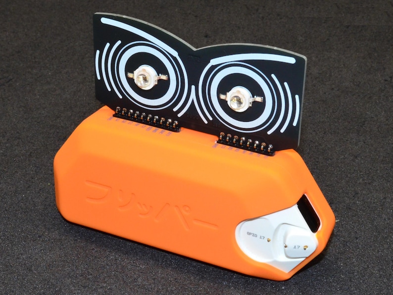 Puede incluir: Un dispositivo electr&oacute;nico naranja con una parte superior negra que presenta dos dise&ntilde;os circulares con c&iacute;rculos conc&eacute;ntricos blancos. El dispositivo tiene una secci&oacute;n blanca con botones y texto japon&eacute;s. El dise&ntilde;o general es moderno y centrado en la tecnolog&iacute;a.