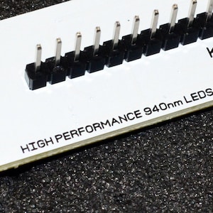 K&ouml;nnte beinhalten: Eine wei&szlig;e Leiterplatte mit einer Reihe schwarzer Stecker und silbernen Pins. Der Text "HIGH PERFORMANCE 940nm LEDs" ist auf der Platine aufgedruckt. Die Platine liegt auf einer dunklen, strukturierten Oberfl&auml;che.