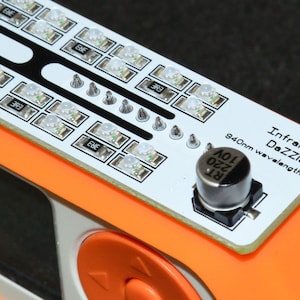 K&ouml;nnte beinhalten: Nahaufnahme eines elektronischen Ger&auml;ts mit einer wei&szlig;en Leiterplatte mit mehreren kleinen LED-Leuchten. Das Ger&auml;t hat einen orangefarbenen Rahmen und den Text "InfraRed DaZZler 940nm wavelength". Eine schwarze zylindrische Komponente ist ebenfalls sichtbar.