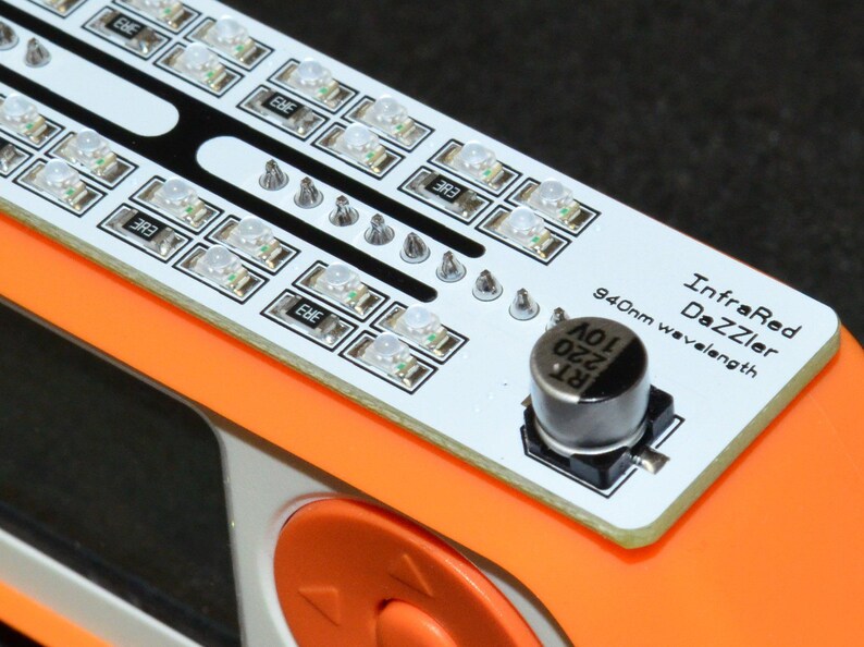 K&ouml;nnte beinhalten: Nahaufnahme eines elektronischen Ger&auml;ts mit einer wei&szlig;en Leiterplatte mit mehreren kleinen LED-Leuchten. Das Ger&auml;t hat einen orangefarbenen Rahmen und den Text "InfraRed DaZZler 940nm wavelength". Eine schwarze zylindrische Komponente ist ebenfalls sichtbar.