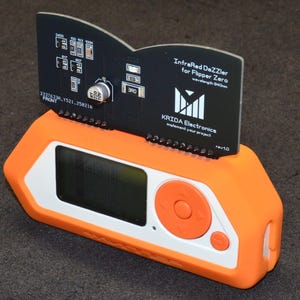 Puede incluir: Un dispositivo electr&oacute;nico naranja y blanco con una pantalla negra y botones. Una placa de circuito negro est&aacute; insertada en el dispositivo, mostrando el texto "InfraRed DaZZler for Flipper Zero" y el logotipo de KRIDA Electronics.